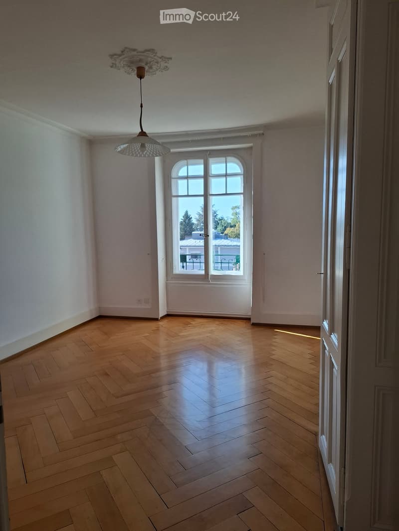 Empty room with wooden herringbone flooring, white walls, and a large window with a view of the outside. A decorative ceiling light fixture is visible.