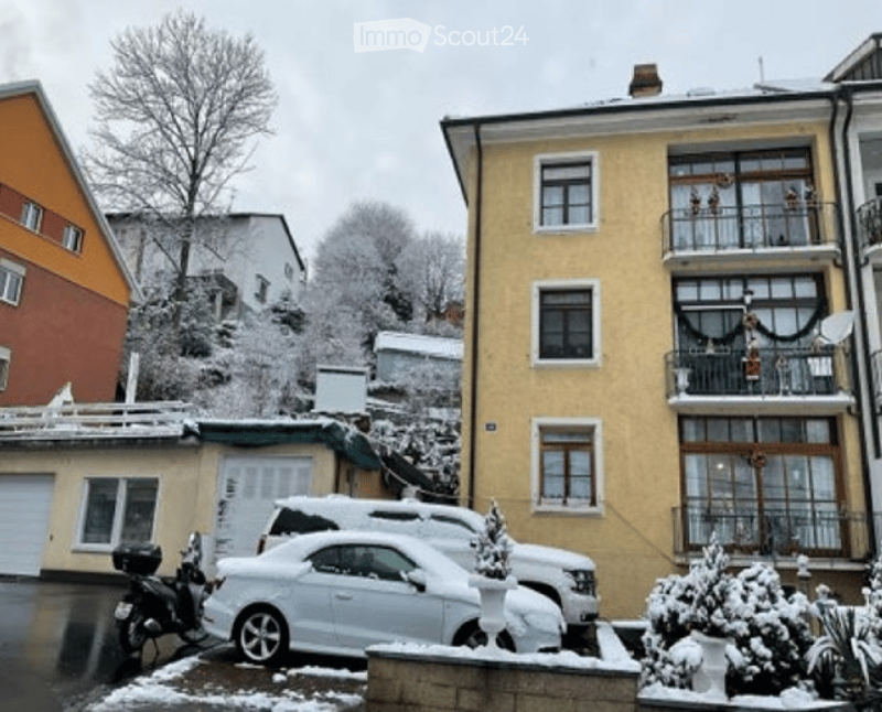 Multi-story residential building with balconies, snow-covered ground, parked cars, and small garden area.