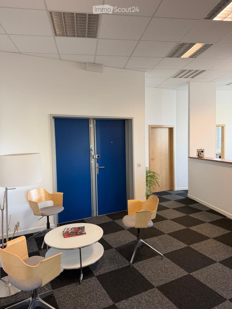 Modern office lounge area with light wood chairs, a round white table, and a floor lamp. The space features a checkered carpet and white walls.