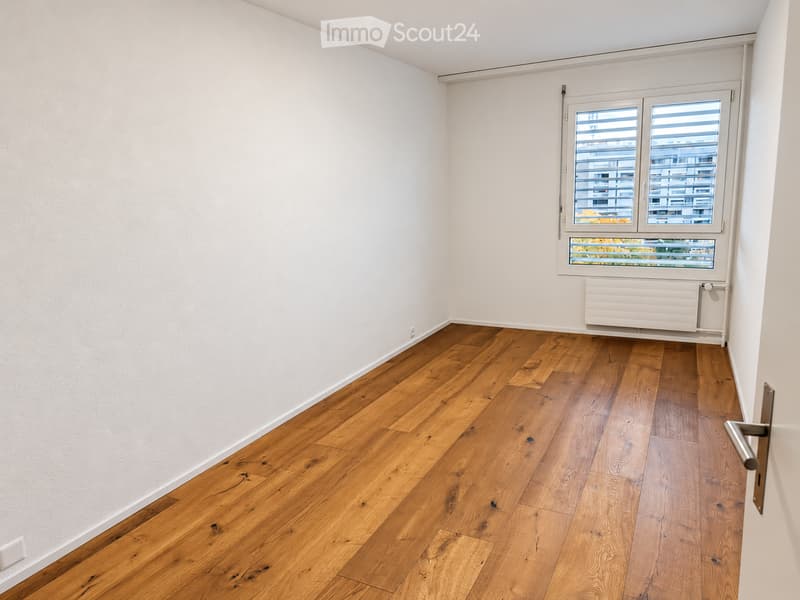 Empty room with light wooden flooring and white walls, featuring a large window with blinds and a radiator below it.