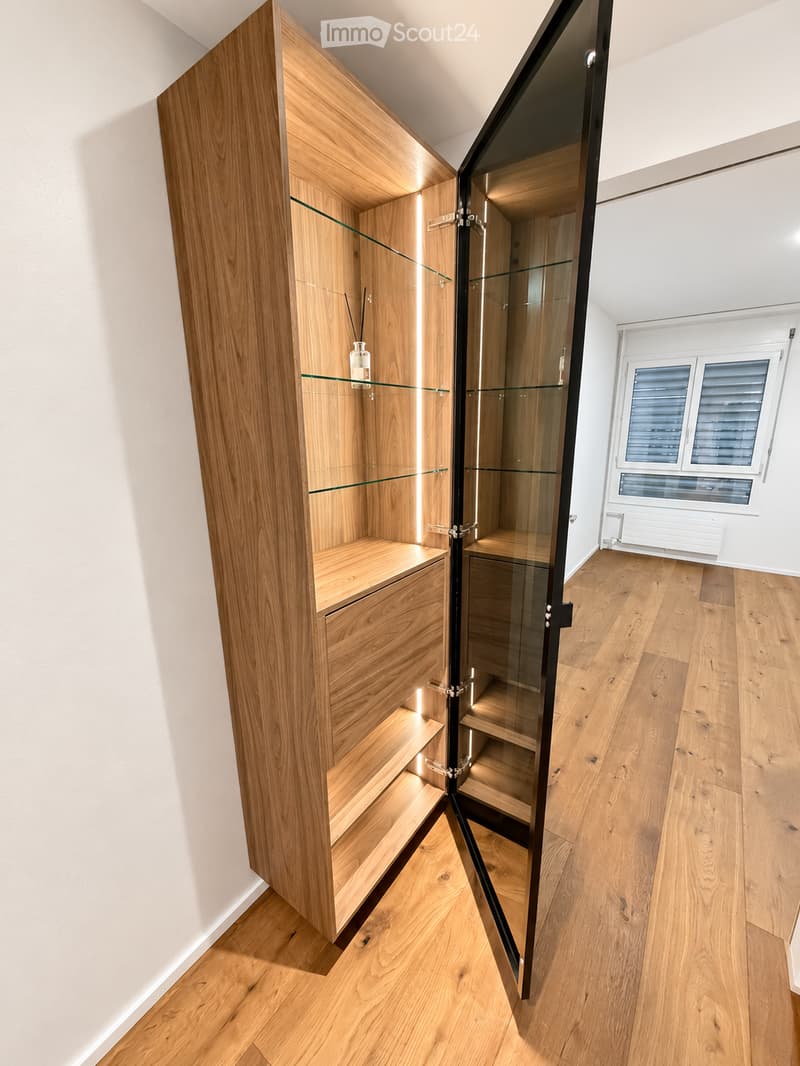 A modern, minimalist wooden cabinet with glass doors and shelves, featuring a single decorative bottle on one of the shelves. The room has light wooden flooring and white walls.