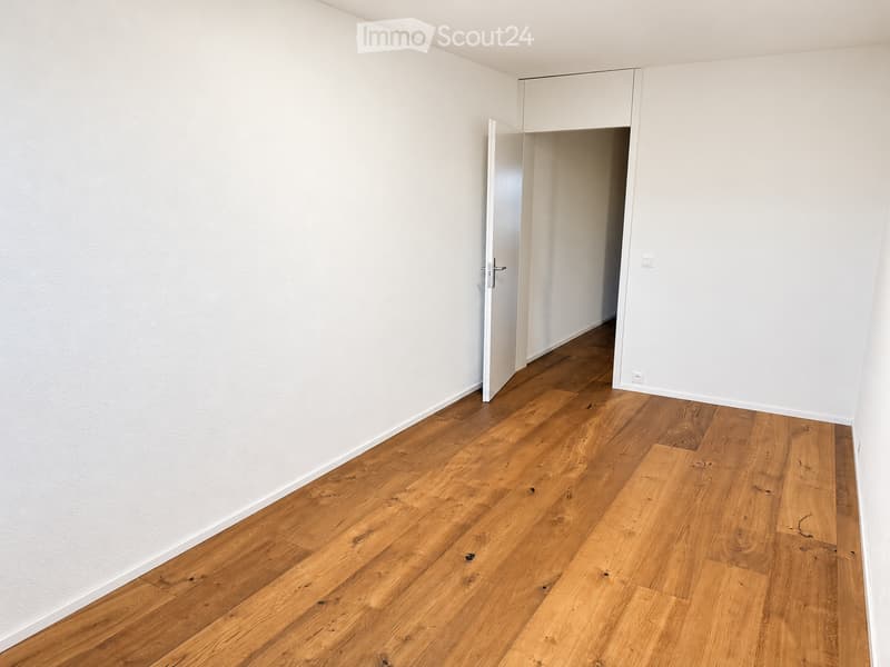 Empty room with light wooden flooring and white walls, featuring an open doorway leading to another room.