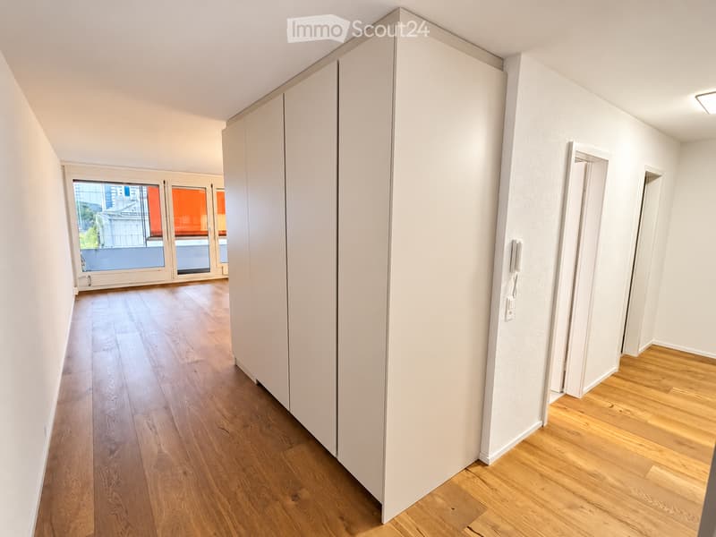 A clean, modern hallway with light wooden flooring, white walls, and a large sliding glass door leading to a balcony area with city views.