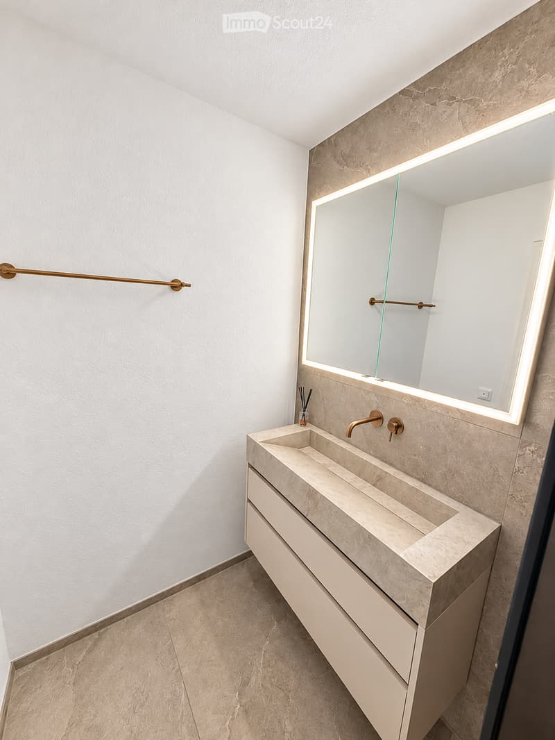 Modern bathroom with a large, illuminated mirror above a stone sink, featuring a sleek faucet and a built-in storage cabinet.