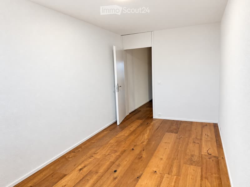 Empty hallway with light wooden flooring and white walls, featuring an open door leading to another room.