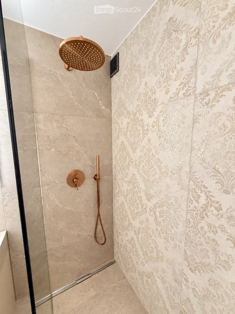 Modern bathroom shower area with beige marble tiles, decorative floral wall pattern, and a sleek copper showerhead and handheld sprayer.