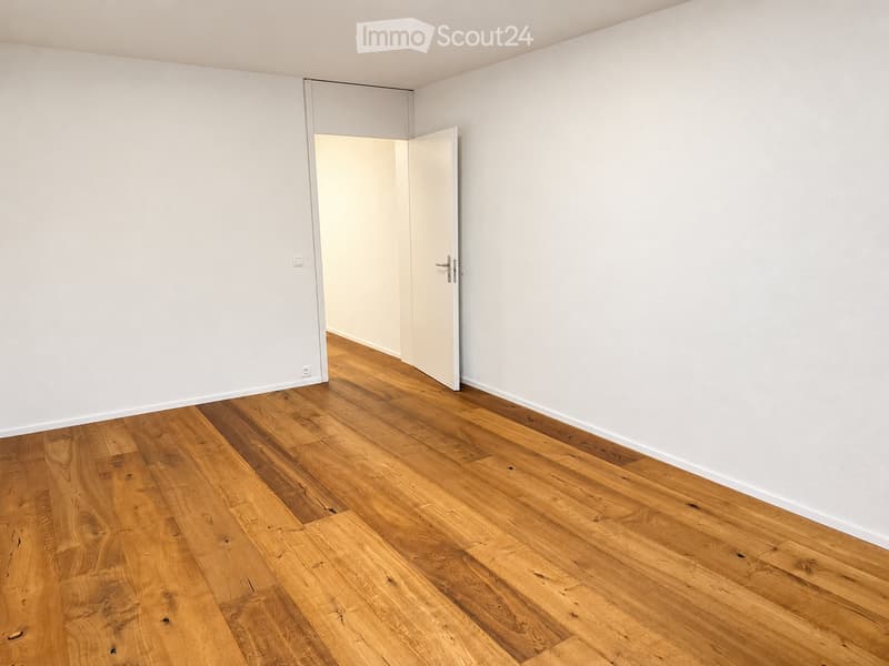 Empty room with light wooden flooring and white walls, featuring an open doorway leading to another space.