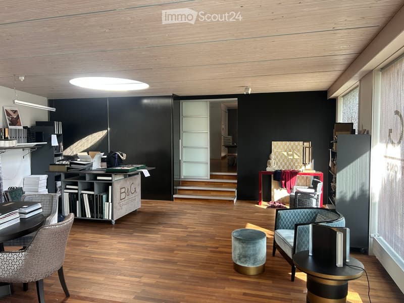Modern, well-lit room with wooden flooring, a large round table, chairs, a shelf with books, a small sofa, a round stool, and a lamp. The room features a sleek, contemporary design with dark walls and large windows.