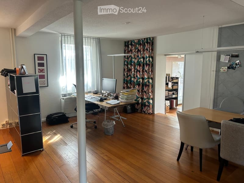 A furnished room with wooden flooring, a large window with curtains, a desk with a computer, a small fridge, and a meeting area with a table and chairs.