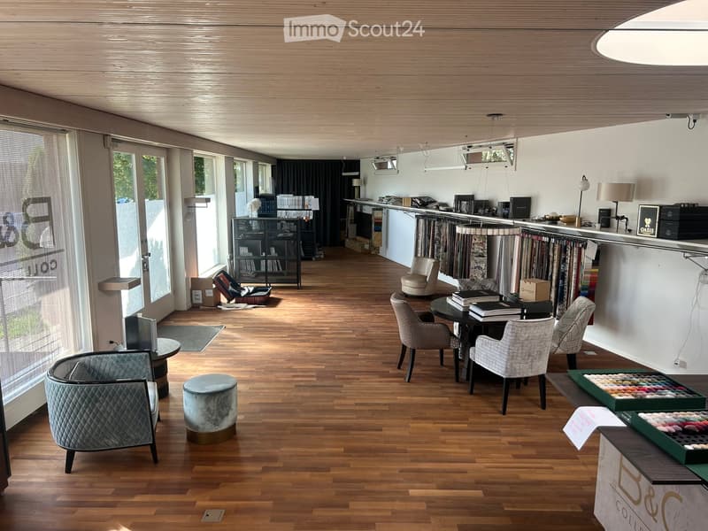 A spacious, well-lit interior with wooden flooring, modern furniture including sofas, chairs, a round table, and a stool, along with shelves and display items.
