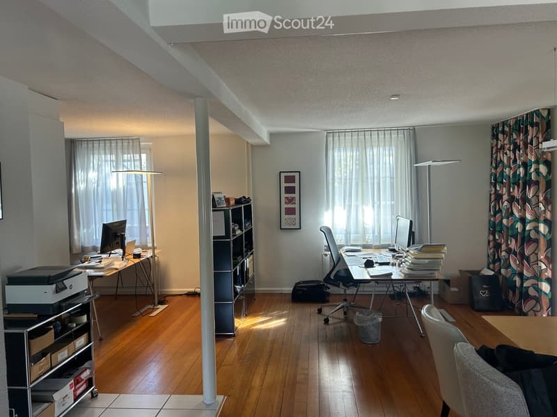 A modern, well-lit office space with wooden flooring, large windows, and multiple desks equipped with computers, chairs, and office supplies.