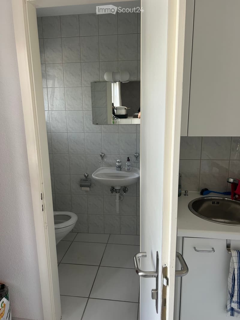 Modern bathroom with grey tiled walls and floor, white cabinetry, a toilet, a sink with a mirror above it, and a towel hanging on the cabinet.
