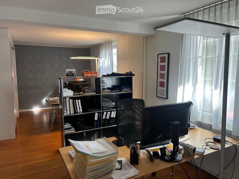 Well-lit office space with wooden flooring, large window with white curtains, desk with computer setup, bookshelf, and various office supplies.