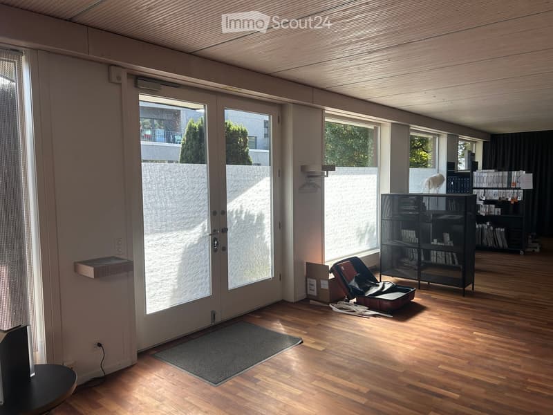 A modern, well-lit room with wooden flooring, large windows, and a door leading outside. Visible items include a small table, a suitcase, shelves with books, and a chair.