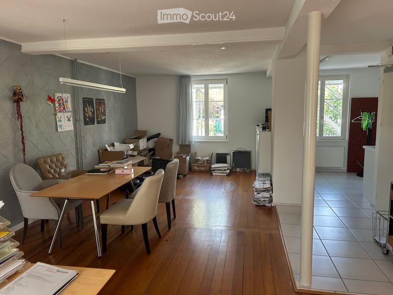A furnished room with a wooden floor, a large table with chairs, a lamp, a refrigerator, and a radiator visible in the background.