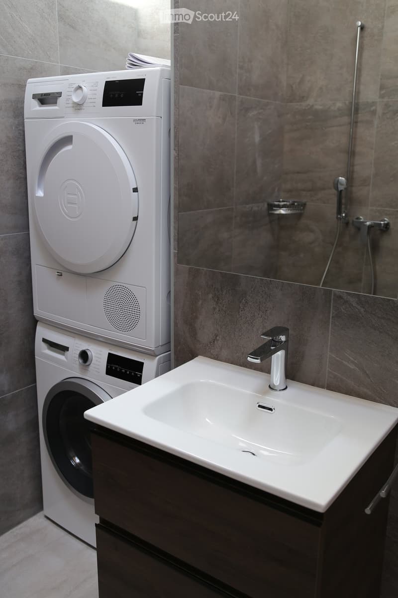 Modern bathroom with a white sink, sleek faucet, and a stacked washing machine and dryer unit. The shower area features a glass enclosure with a modern design.