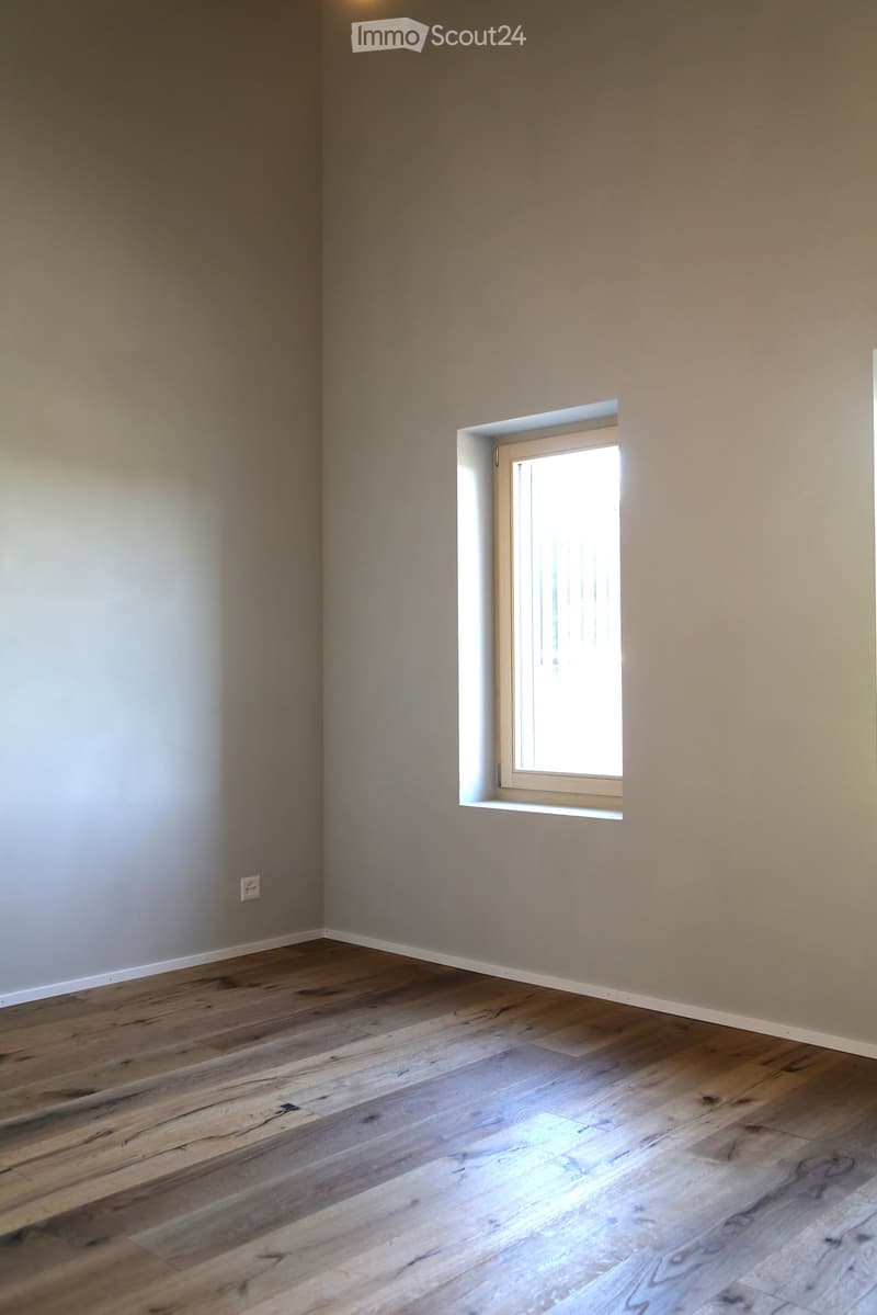Empty room with wooden flooring and light-colored walls, featuring a small window with white frame and a visible electrical outlet on the left wall.