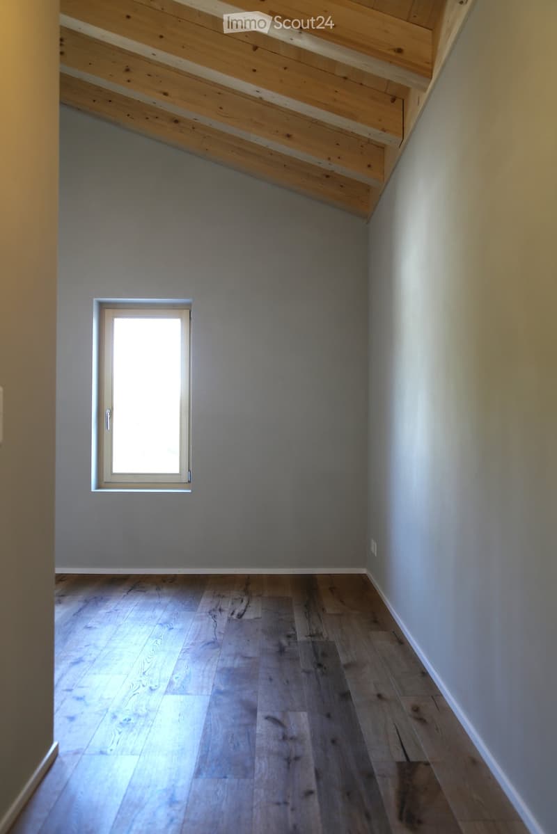 Empty hallway with wooden flooring, light grey walls, and a small window allowing natural light to enter.