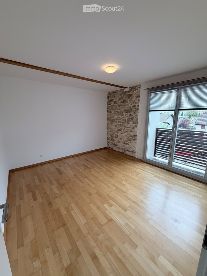 Empty room with wooden flooring, white walls, and a large window with blinds. Exposed stone wall visible on one side.