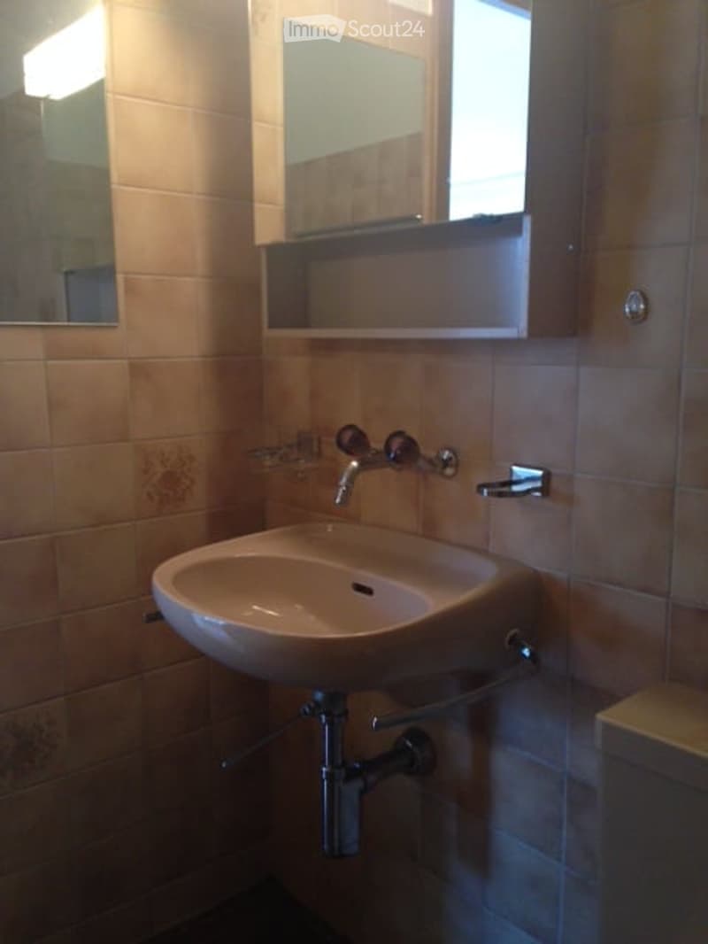 A small bathroom with a white sink, a chrome faucet, a mirror above the sink, and a wooden cabinet with a shelf. The walls are tiled in a light brown color.