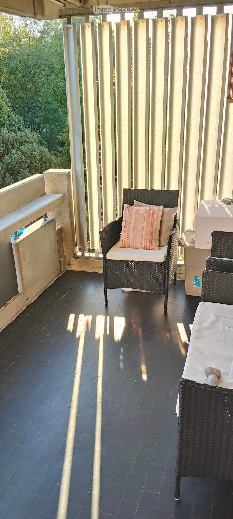Balcony with dark tiled flooring, wicker chairs, and a small table, featuring a view of greenery through vertical slats.