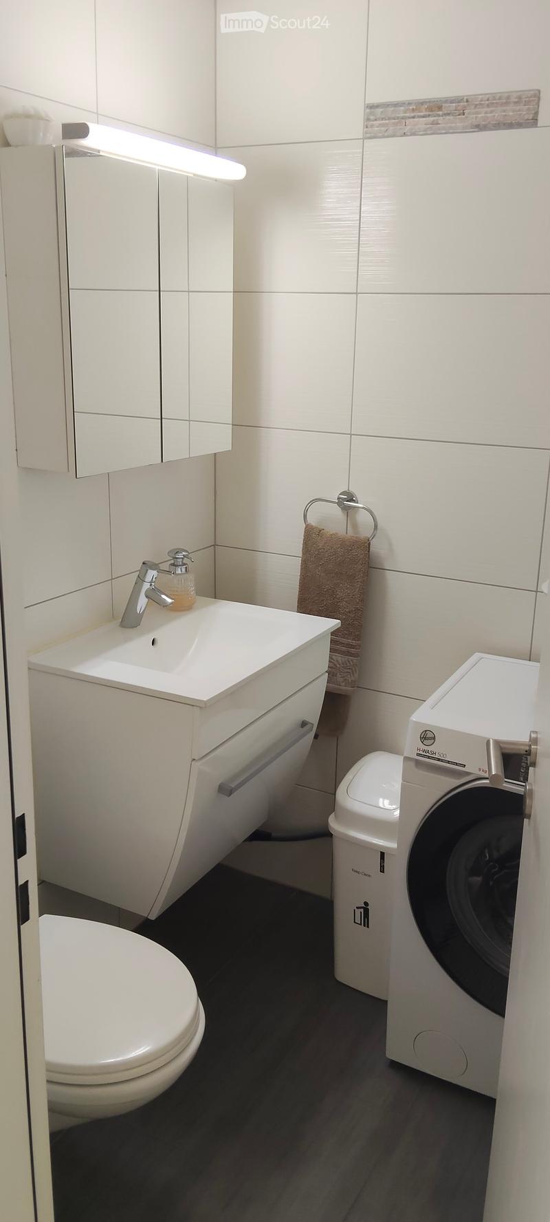Modern bathroom with white tiles, a sink with a modern faucet, a toilet, a mirror cabinet, a towel, and a washing machine.