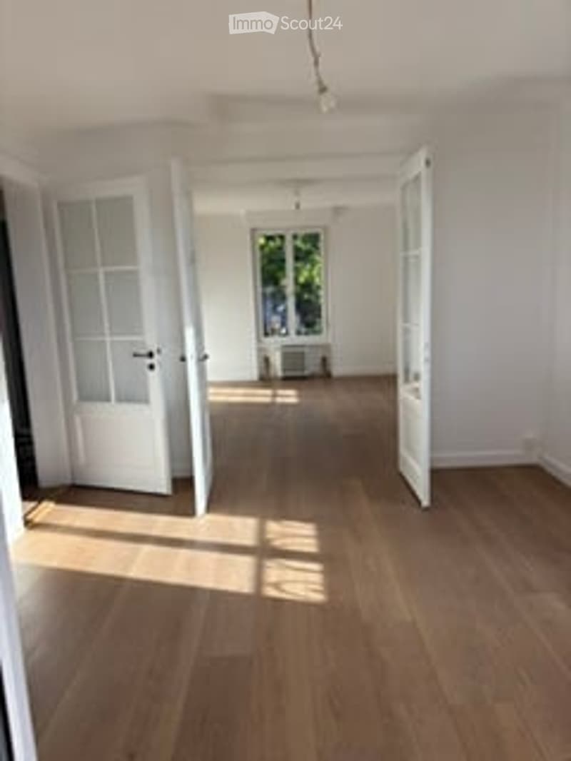 Empty room with wooden flooring, white walls, and large open doors leading to another space.