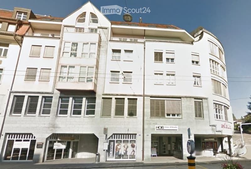 Multi-story building with white facade, multiple windows, and balconies. Ground floor has commercial storefronts with signage.