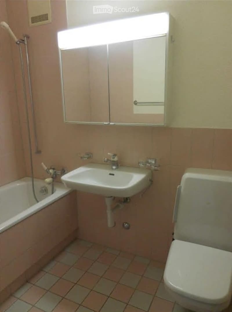 Bathroom with a bathtub, sink, toilet, and a mirror with built-in cabinets above it. The walls are tiled in a warm, peach color, and the floor has a mix of peach and gray tiles.