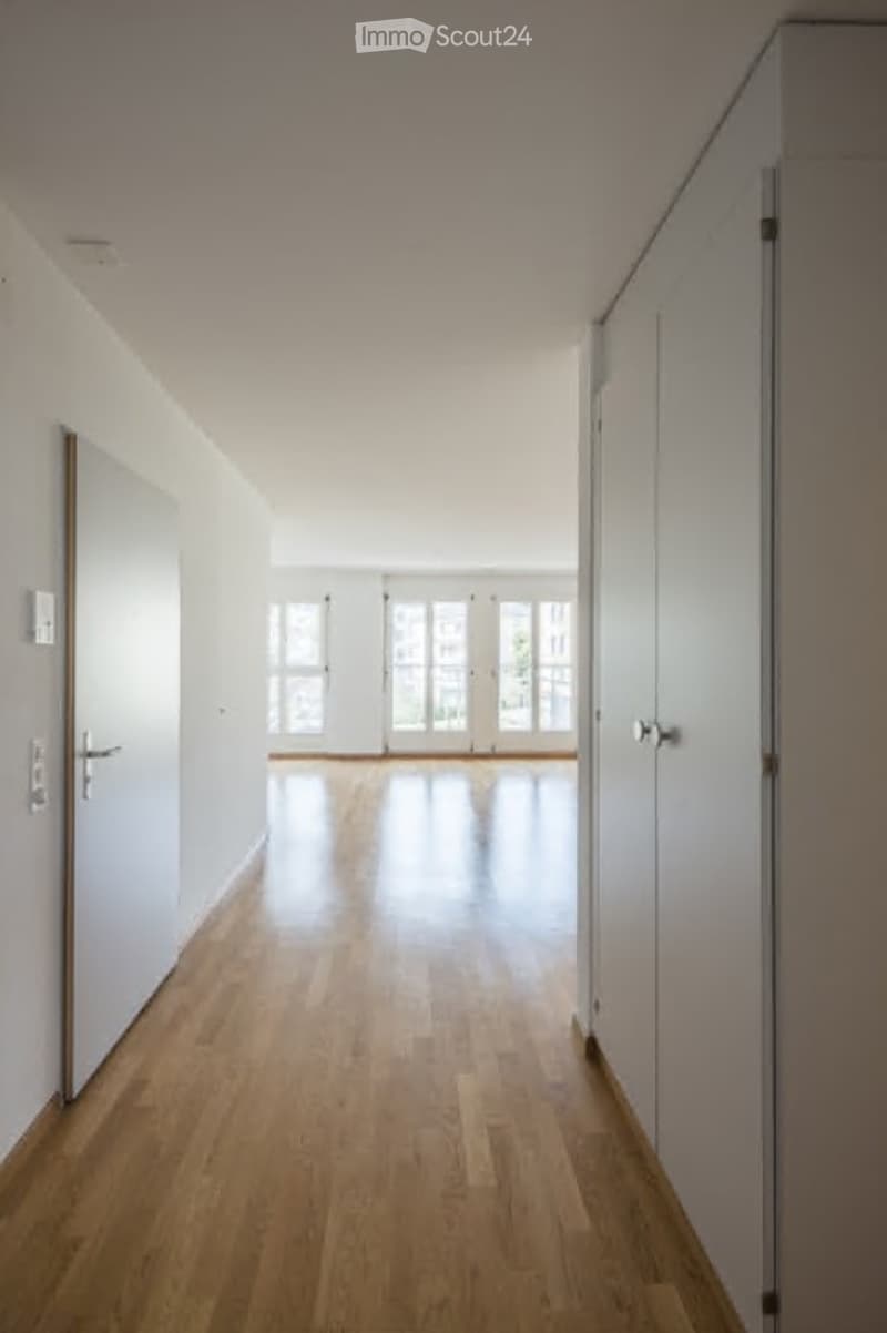 Empty hallway with light wooden flooring, white walls, and large windows at the end.