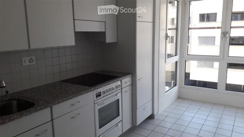 Modern kitchen with white cabinets, granite countertop, built-in oven, and large windows offering a view of the building outside.
