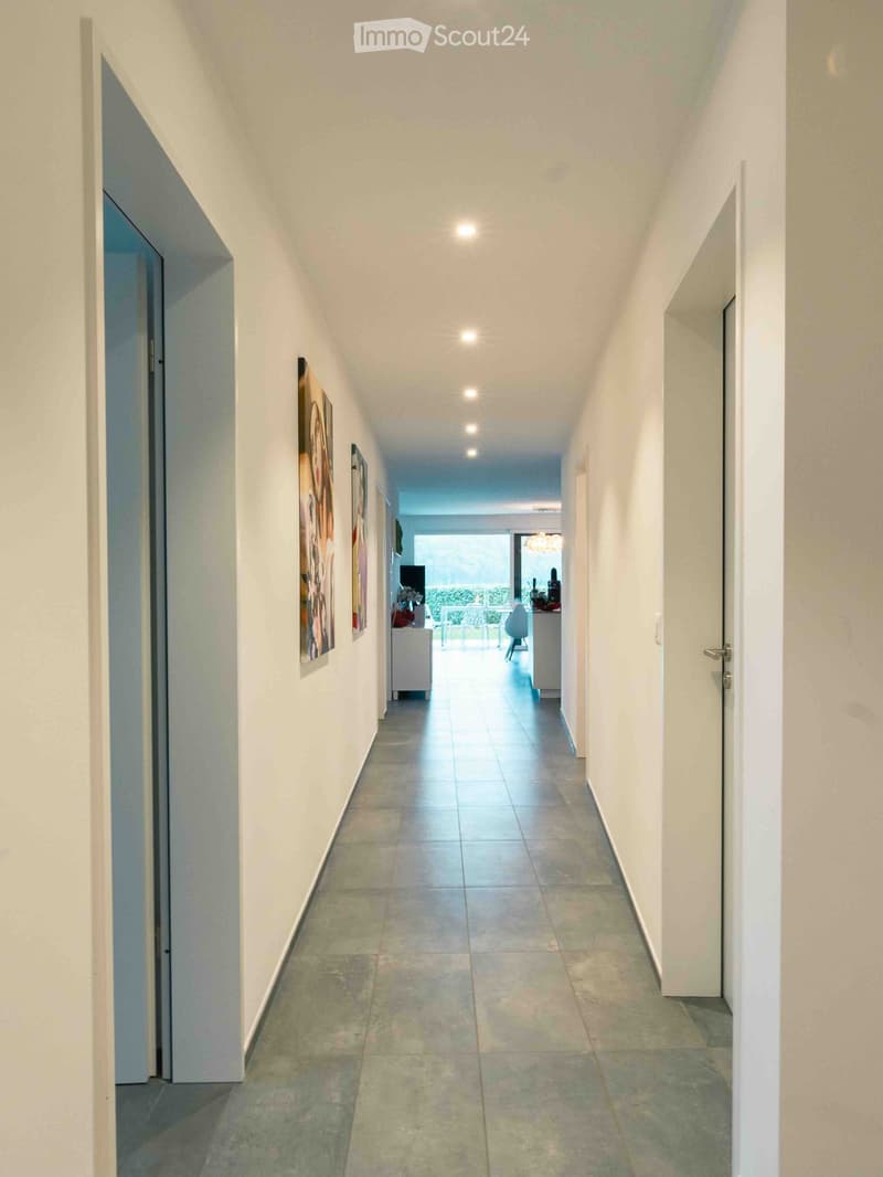 A clean, well-lit hallway with tiled flooring, white walls, and several doors leading to different rooms. Artwork is displayed on one wall, and the corridor leads to a brightly lit room with a view of the outdoors.