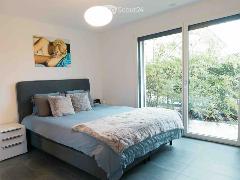Modern bedroom with a large bed, nightstand, and artwork on the wall. Large sliding glass doors lead to a lush outdoor area.