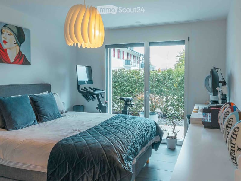 Modern bedroom with a large bed, exercise bike, pendant light, and a view of greenery through large windows.