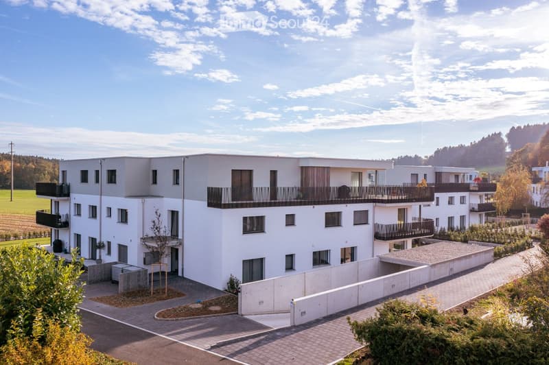 Modern multi-story residential building with white facade, multiple balconies, and surrounding greenery.