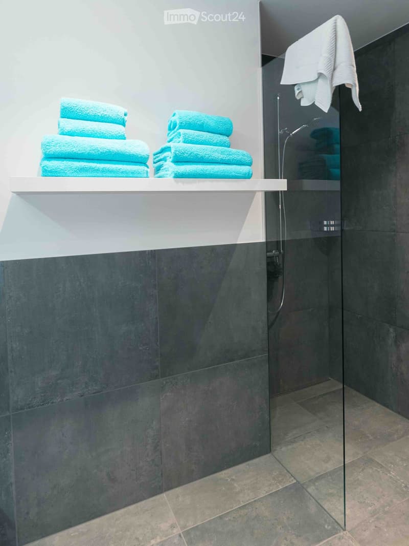Modern bathroom with dark grey tiles, glass shower enclosure, and neatly stacked blue towels on a white shelf.