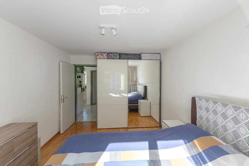 Modern bedroom with a large bed, colorful bedding, a large mirrored wardrobe, a nightstand, and a wooden drawer unit. The room features wooden flooring and white walls.