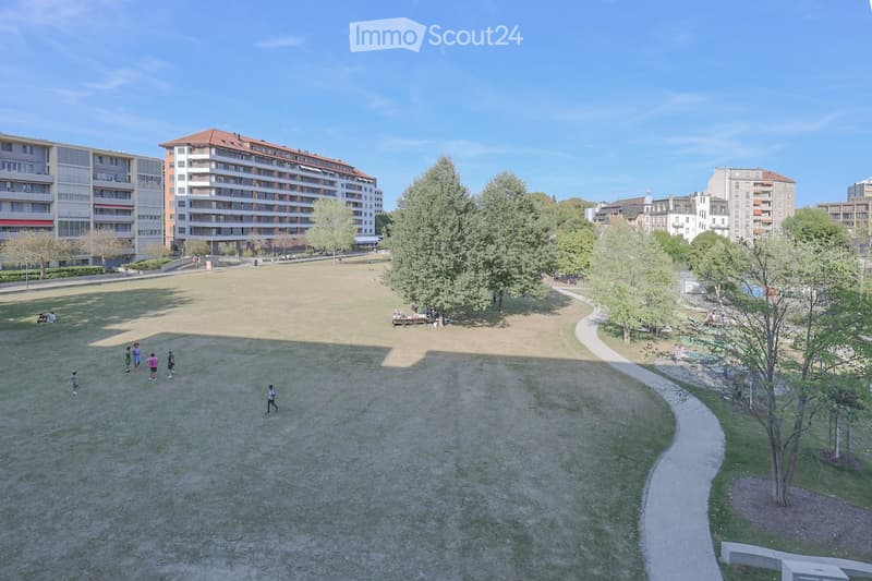 A spacious park area with grassy fields, trees, pathways, and surrounding residential buildings.