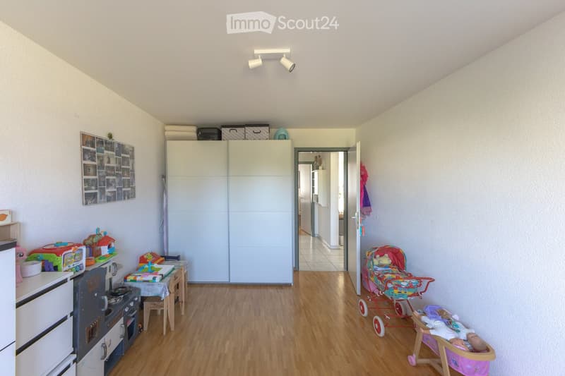 Brightly lit children's room with wooden flooring, a toy stove, a small table with chairs, a large wardrobe, a baby stroller, and a baby crib.