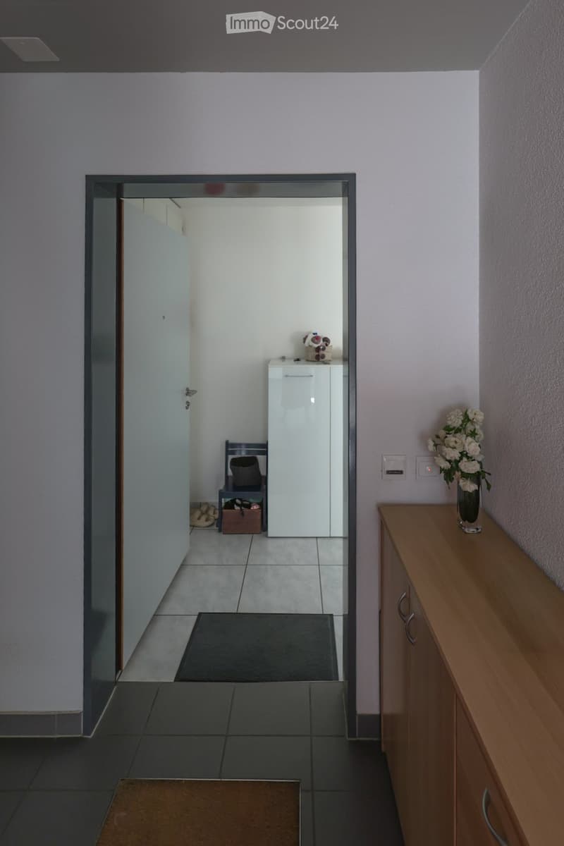 A modern hallway with a tiled floor, light-colored walls, and a wooden console table with a vase of white flowers. A small bench and a white cabinet are visible in the adjacent room.