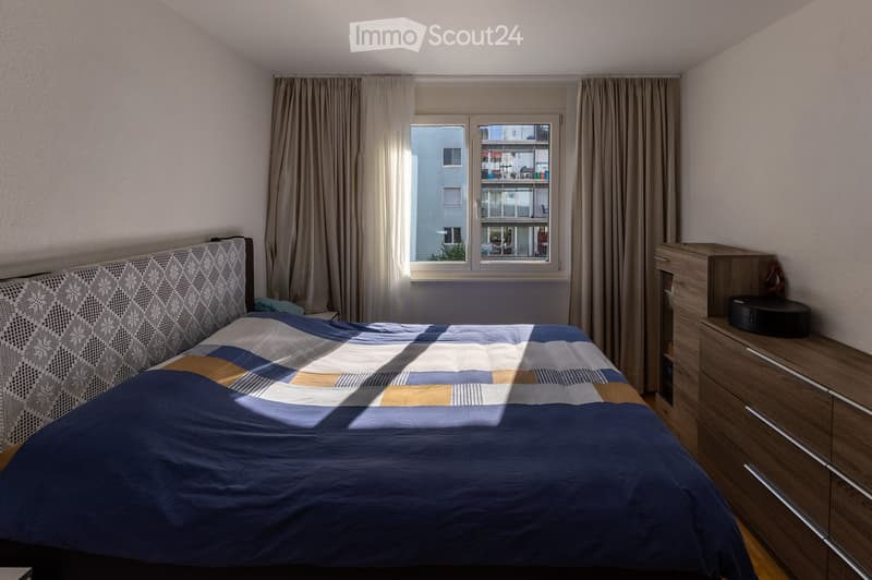 A modern bedroom with a large bed, patterned headboard, and a matching duvet. A wooden wardrobe with drawers is positioned next to the bed, and a window with curtains offers a view of the city.