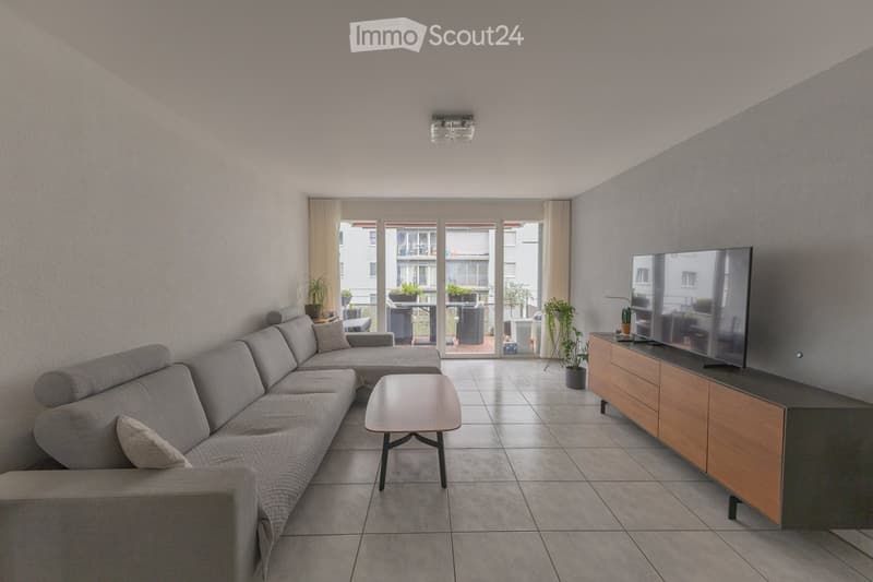 Modern living room with grey sectional sofa, coffee table, TV stand, and potted plants near large windows leading to a balcony.