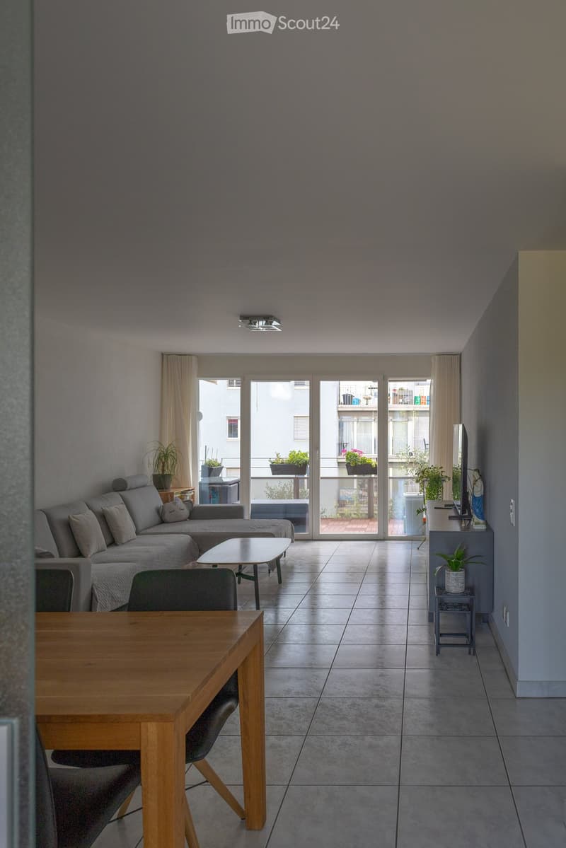 Living room with a sofa, coffee table, dining table, chairs, and plants, leading to a balcony with outdoor seating.