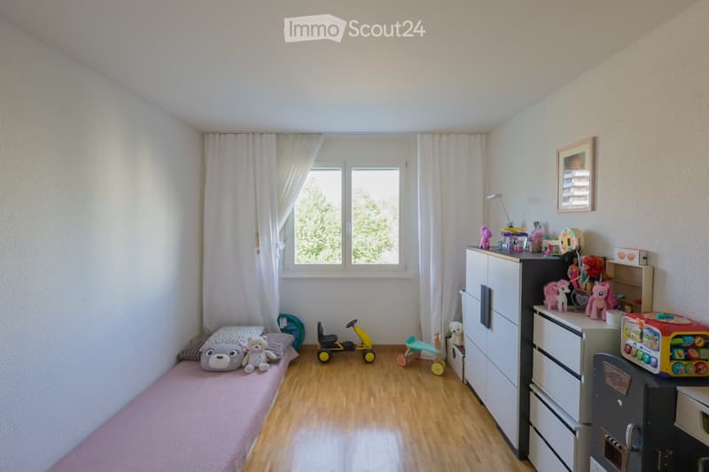 A well-lit children's bedroom with wooden flooring, a small bed with pillows and stuffed animals, a toy car, a toy horse, a toy kitchen set, and various toys scattered around. The room features white walls, large windows with white curtains, and a white w