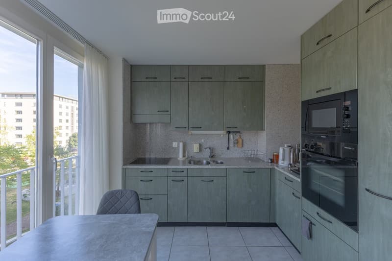 Modern kitchen with grey cabinets, stainless steel appliances, and a large window with white curtains. Includes a microwave, oven, sink, and a small dining area with a chair and table.