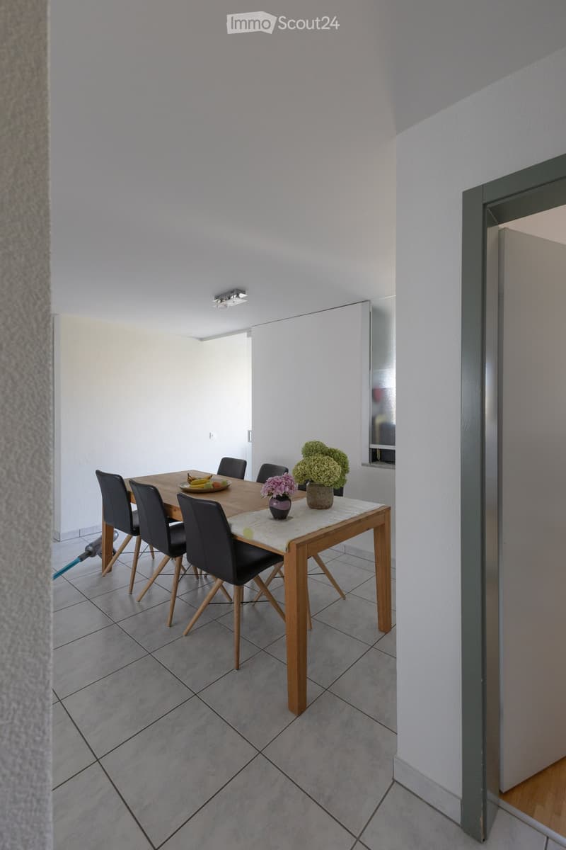 Modern dining room with a wooden dining table, four black chairs, a vase with flowers, and a bowl of fruit on the table. The room features tiled flooring and white walls.