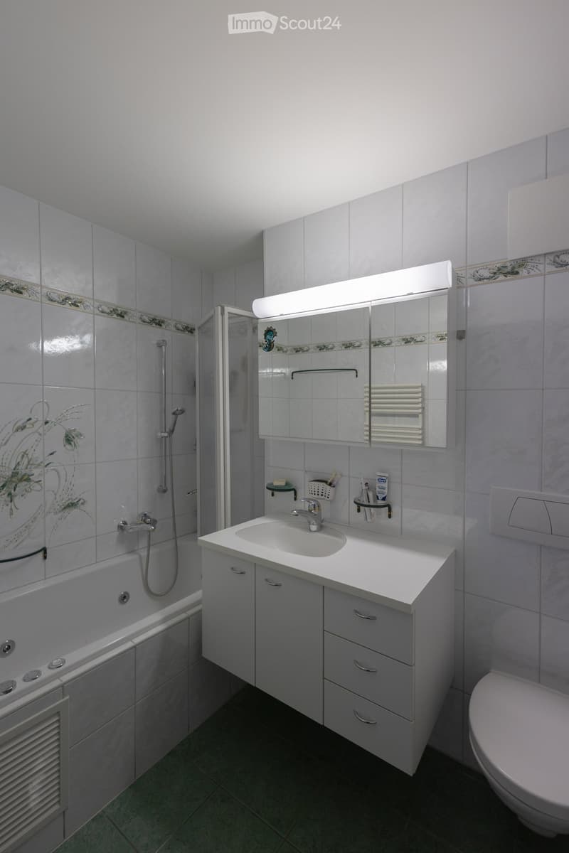 Modern bathroom with white tiles, a sink with a mirror cabinet, a bathtub with a shower, and a toilet. Features include a built-in radiator, a toothbrush holder, and a soap dispenser.