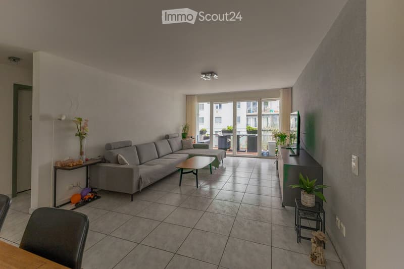 Modern living room with grey sectional sofa, coffee table, TV stand, and balcony access. Features tiled flooring, wall-mounted lamp, and decorative plants.
