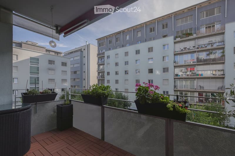 Balcony with wooden decking, metal railing, wicker chair, and several flower pots with plants and flowers.