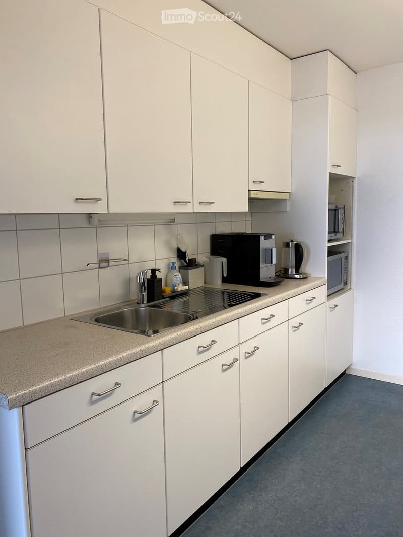Modern kitchen with white cabinets, a stainless steel sink, a coffee maker, a kettle, and a microwave. The countertops are light beige, and the floor is blue.